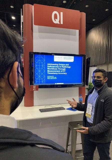 man watching male trainee present about quality improvement in a large exhibit hall with presentation on a tv sized monitor