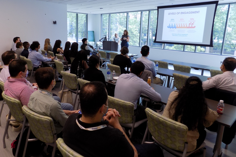 large group of diverse students wearing masks sitting at tables listening to two speakers with windows and a projected image about levels of engagement behind them