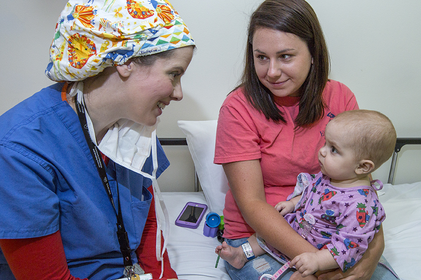 doctor in brightly colored hat and blue shirt smiles at baby which is sitting in its mother's lap