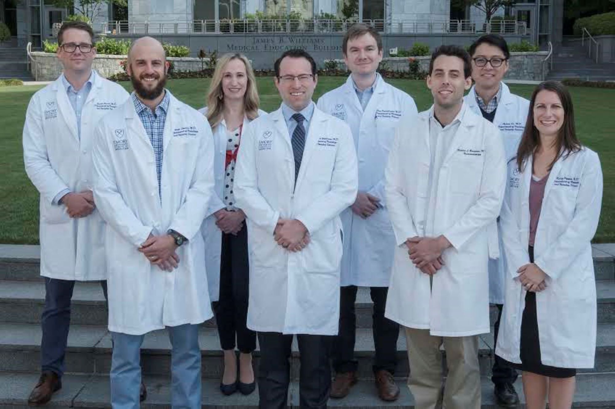 eight people in white doctor coats standing on green lawn in front of grey marble building