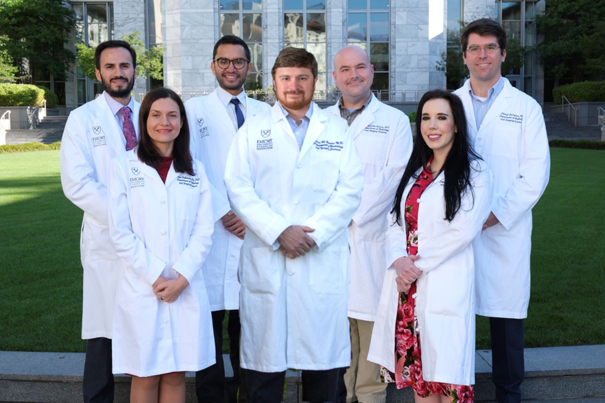 seven people wearing white doctor coats standing on green lawn in front of grey marble building