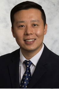 smiling person with short dark hair wearing a dark suit jacket over a white shirt and blue patterned tie