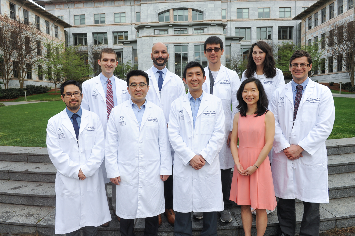 men and women smiling and wearing white doctor coats standing in front of the grey and peach marbled two story School of Medicine building with a green lawn