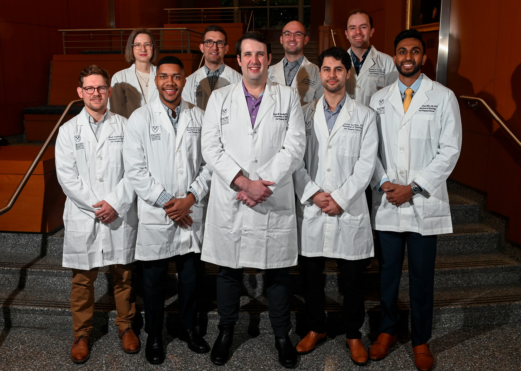 nine smiling people wearing white doctor coats standing in two rows