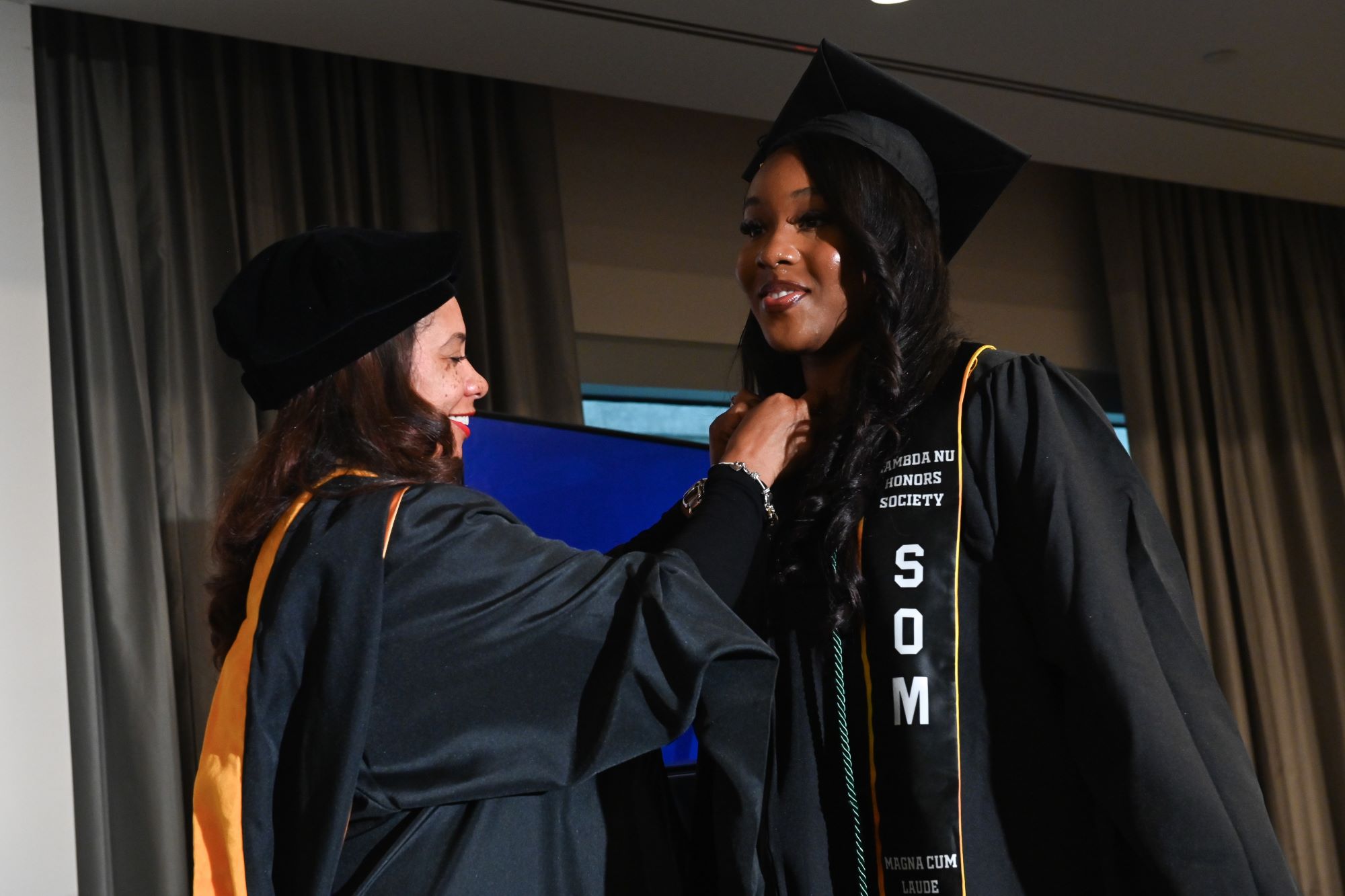 smiling person with lambda nu sash over graduation gown receiving a pin