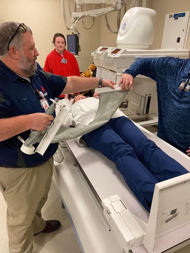 person placing protective covering over person who is laying on the table of an imaging machine with another person helping