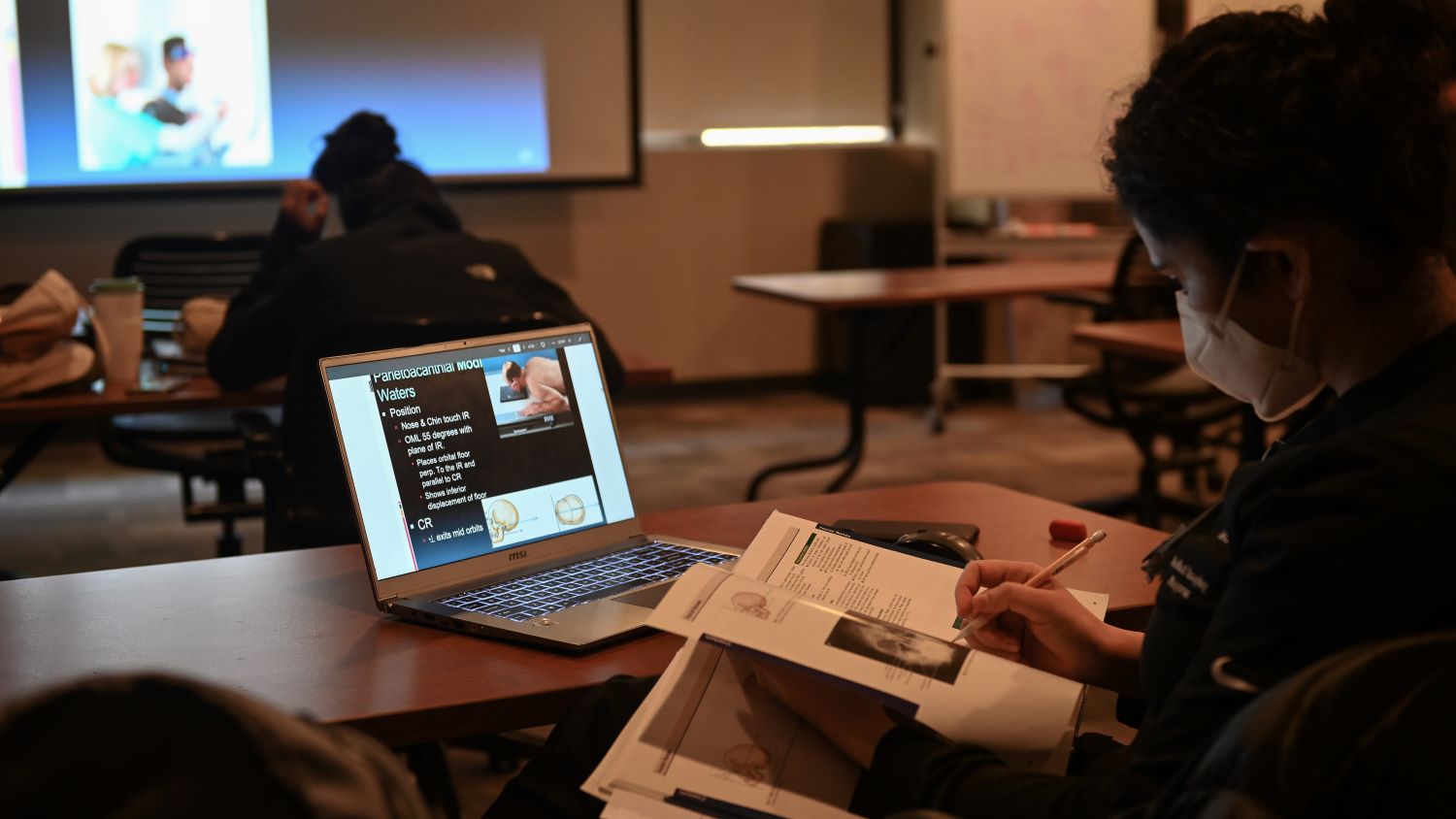 studentin darkened room looking at computer screen with larger projection on screen in front of room