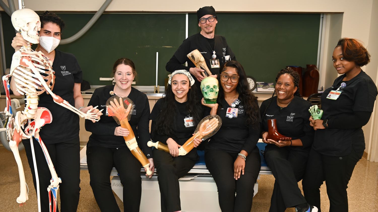 young women and man smiling and each holding a plastic arm or leg or foot or hand or skull and one standing with a full size skeleton holding its hand