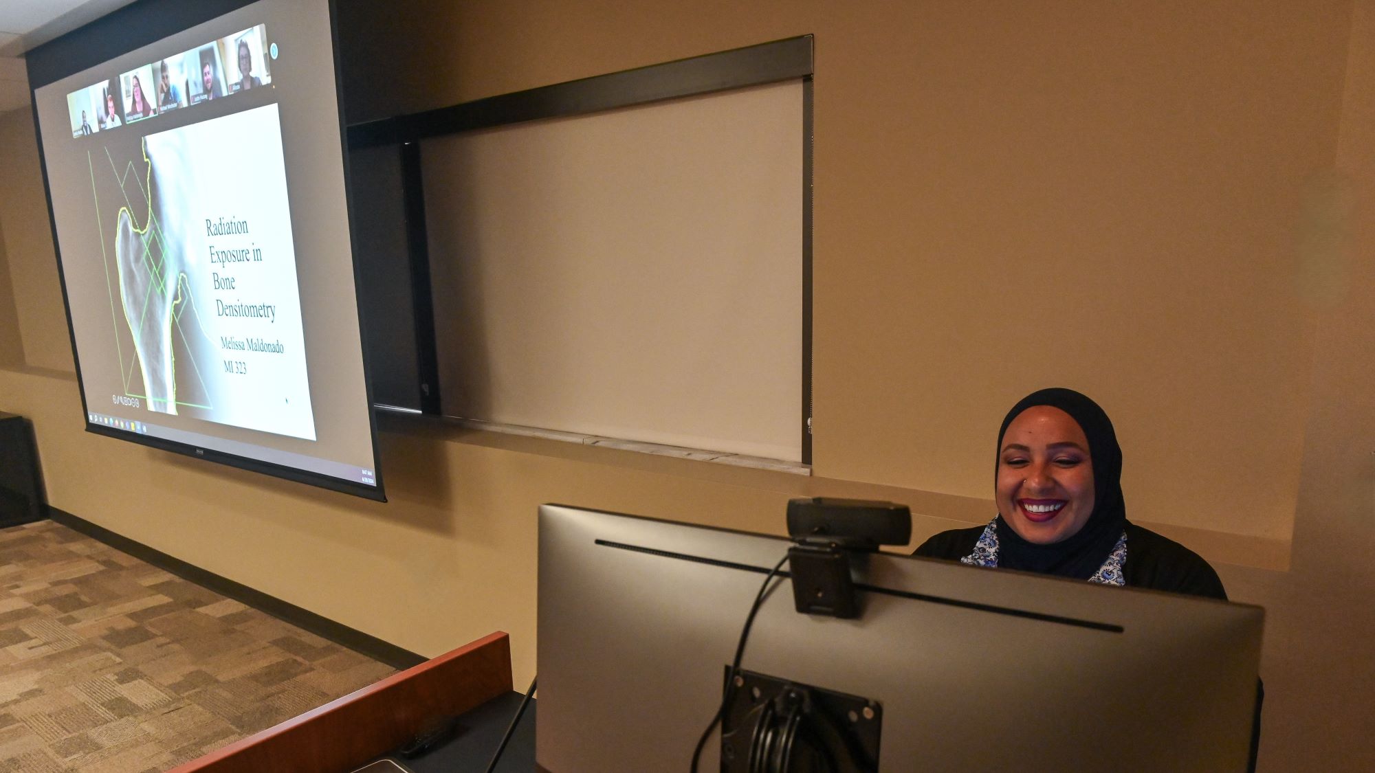 smiling person sitting at lecturn with computer monitor in front of her and a large screen with the images of all her students on the wall