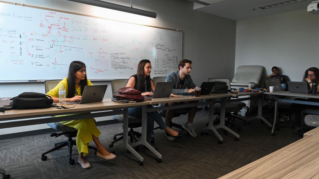 people sitting at tables in classroom working at computers with code written on whiteboard on wall