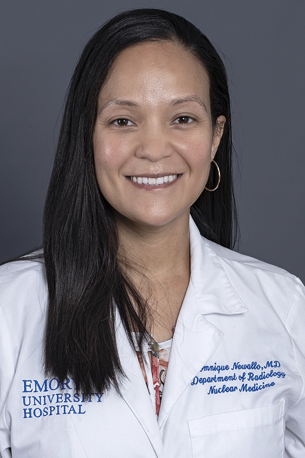 smiling woman with long black hair wearing a white doctor coat embroidered with the name  Domniqe Newallo MD and the coat is over a floral topover